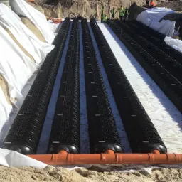 Montaż drenażu rozsączającego z rur drenarskich w wykopie wyłożonym geowłókniną, widoczni pracownicy w odblaskowych kamizelkach i maszyny budowlane w tle.