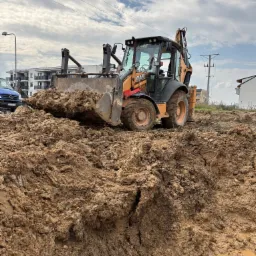 Żółto-czarna koparko-ładowarka CASE z pełną łyżką mokrej ziemi na placu budowy, w tle budynki mieszkalne i pochmurne niebo.