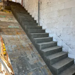 Surowe, betonowe schody wewnętrzne w trakcie budowy, z widocznym szalunkiem i ścianą z bloczków betonowych. Ślady po zaprawie na ścianie.