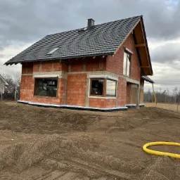 Surowy, dwukondygnacyjny dom z czerwonej cegły w Jeleniej Górze, z ciemnym dachem i zamontowanymi oknami, w trakcie budowy, otoczony ziemią i elementami ogrodzenia.