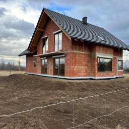 Firma Żelbet - Surowy stan domu z czerwonej cegły z oknami i czarnym dachem, otoczony świeżo przekopaną ziemią i ogrodzeniem z drutu.