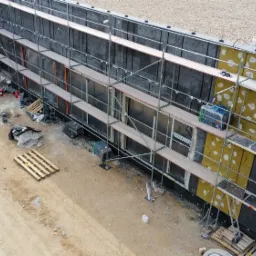 Widok z góry na budynek w trakcie ocieplania, z widocznymi rusztowaniami, częściowo ocieploną ścianą wełną mineralną oraz ekipą budowlaną w oddali.