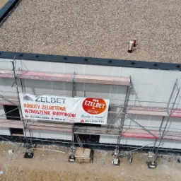 Wysoki kąt widzenia na budynek w trakcie budowy z szarymi ścianami, płaskim dachem pokrytym żwirem i rusztowaniem; baner reklamowy firmy budowlanej zawieszony na rusztowaniu.