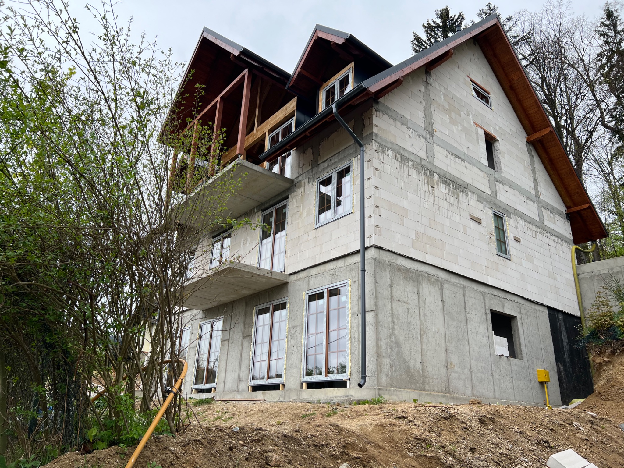 Dwupiętrowy dom w trakcie budowy, widoczne ściany z betonu komórkowego, drewniana konstrukcja dachu, wstawione okna, teren wokół w trakcie prac ziemnych, żółty wąż leży na ziemi.