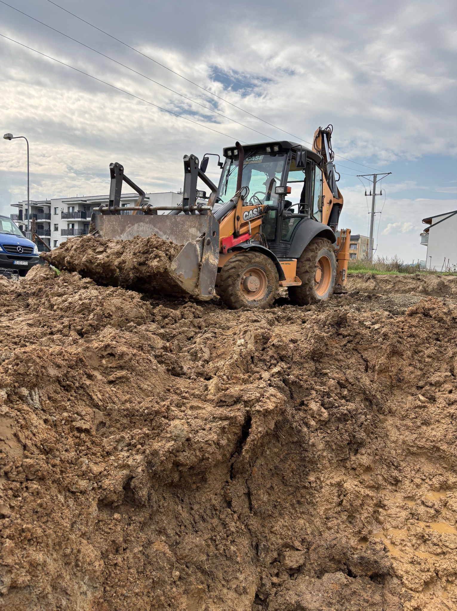 Żółto-czarna koparko-ładowarka CASE z pełną łyżką mokrej ziemi na placu budowy, w tle budynki mieszkalne i pochmurne niebo.