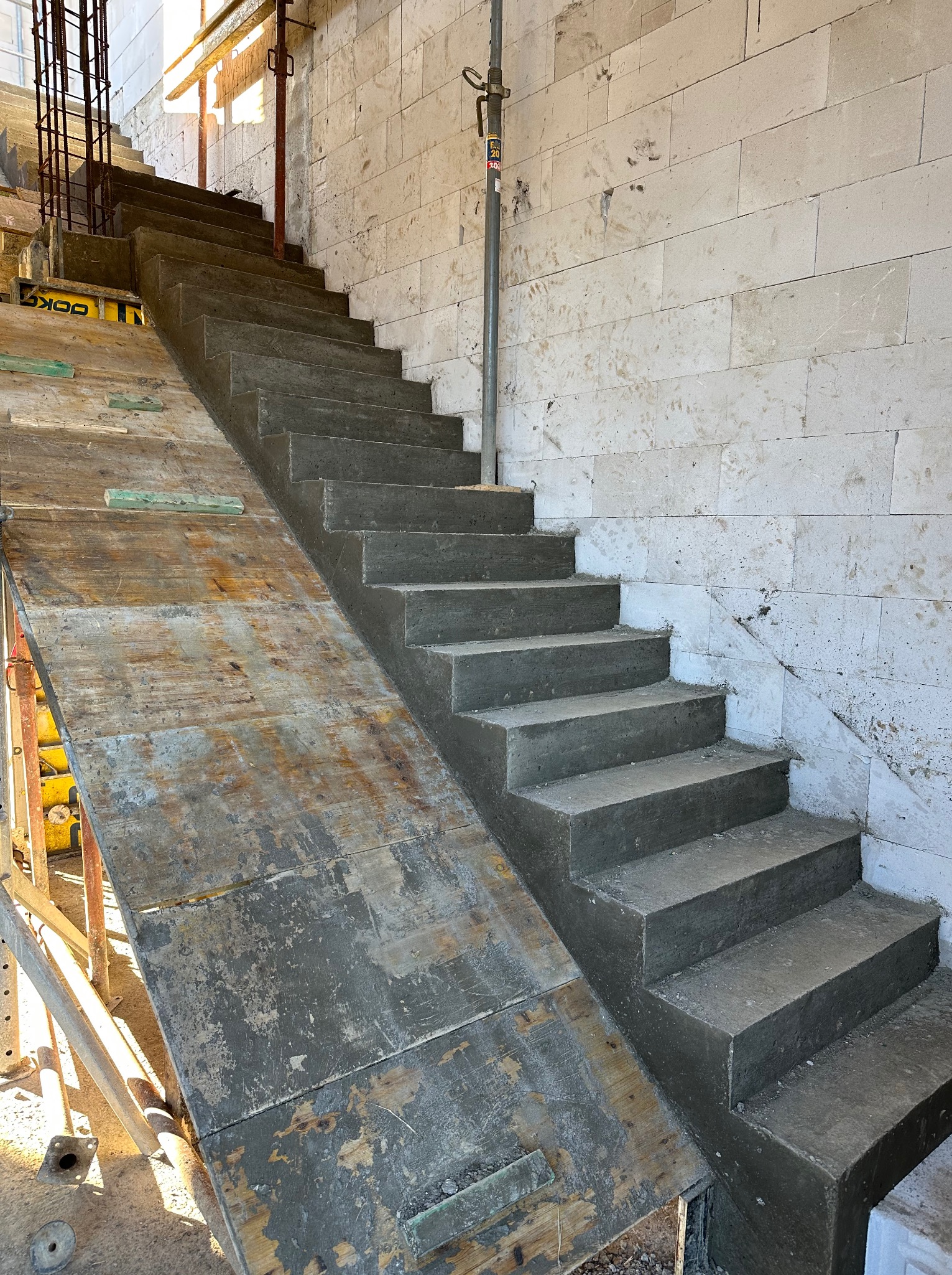 Surowe, betonowe schody wewnętrzne w trakcie budowy, z widocznym szalunkiem i ścianą z bloczków betonowych. Ślady po zaprawie na ścianie.