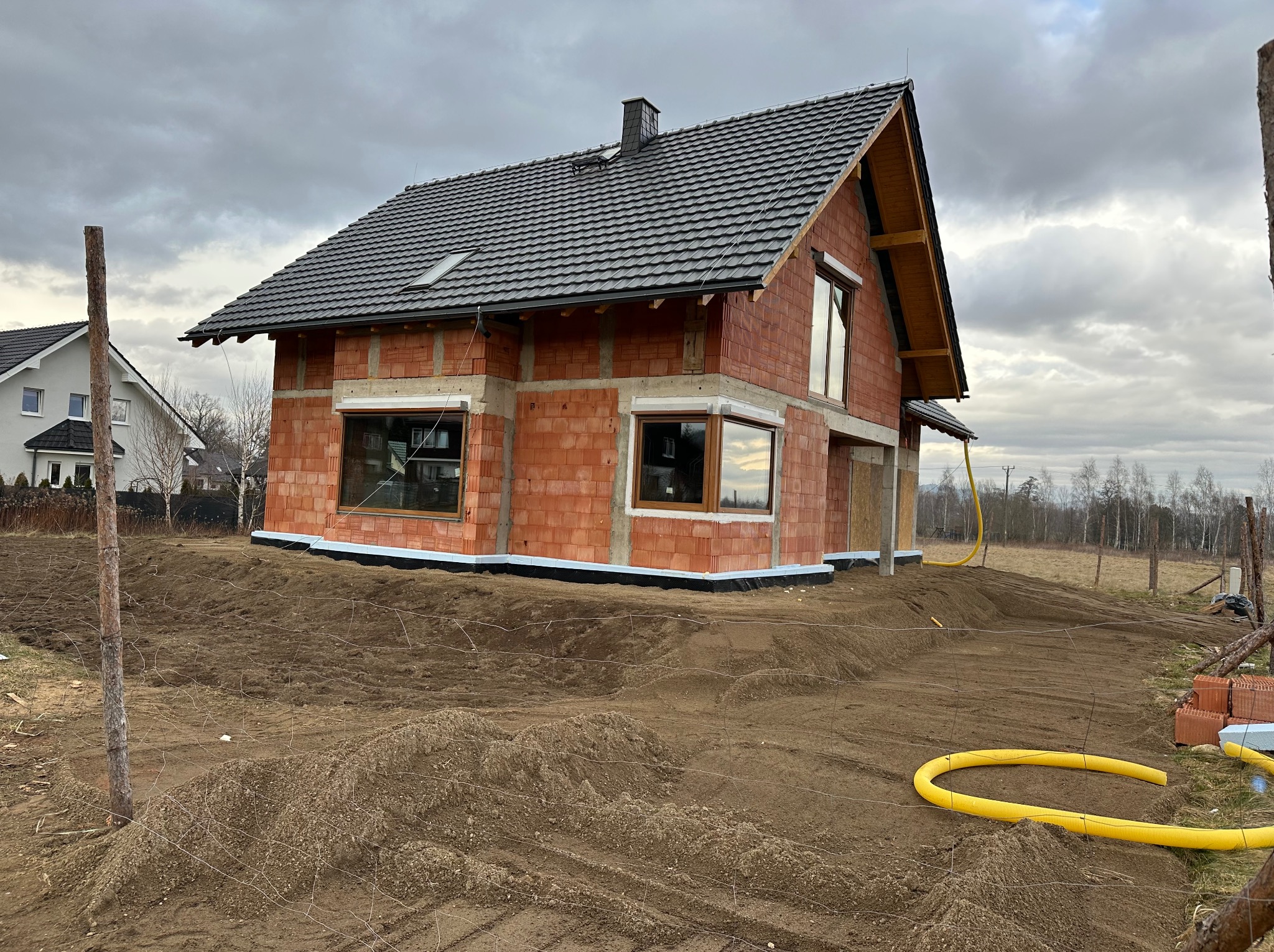 Surowy, dwukondygnacyjny dom z czerwonej cegły w Jeleniej Górze, z ciemnym dachem i zamontowanymi oknami, w trakcie budowy, otoczony ziemią i elementami ogrodzenia.