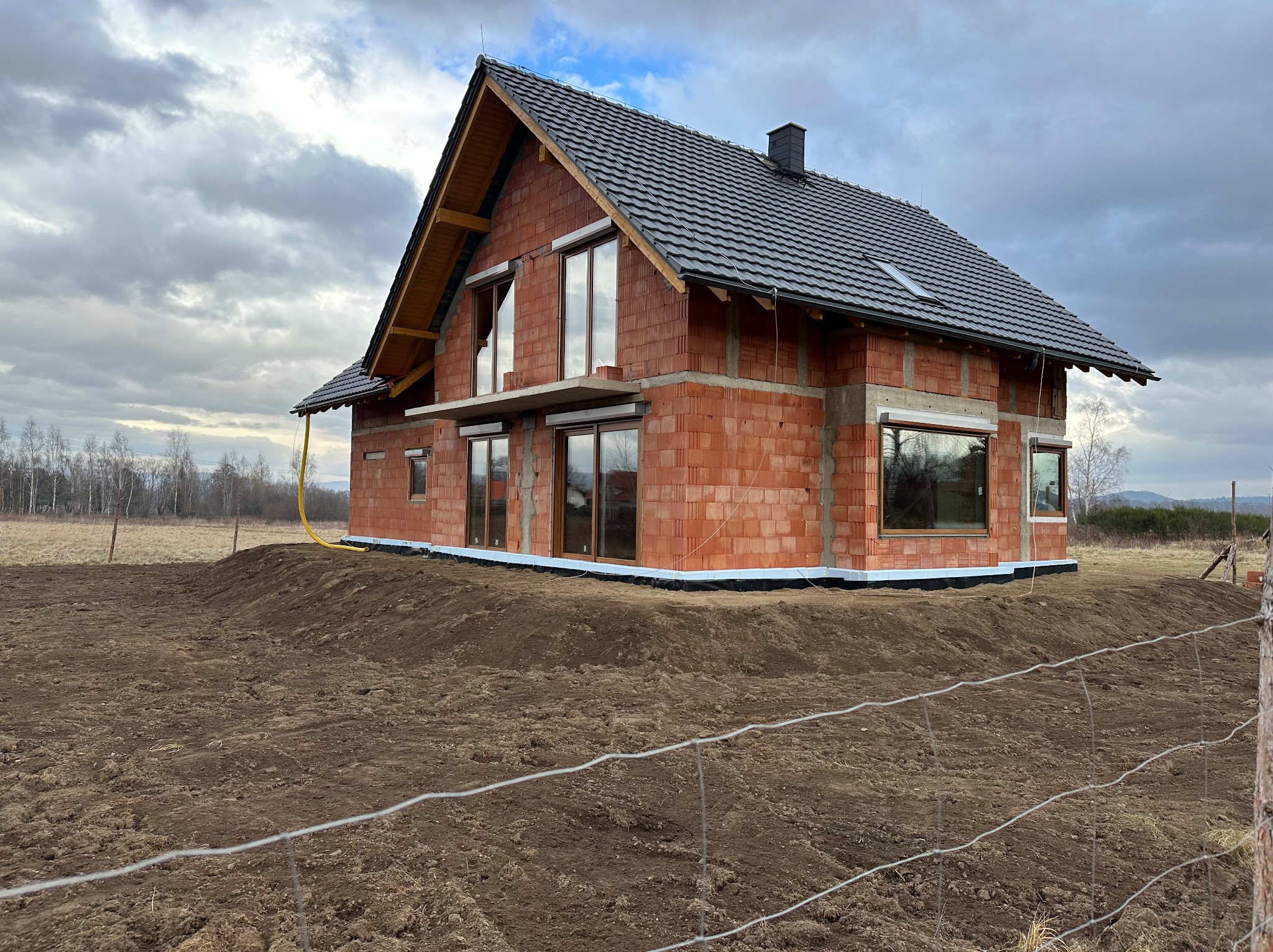 Surowy stan domu z czerwonej cegły z oknami i czarnym dachem, otoczony świeżo przekopaną ziemią i ogrodzeniem z drutu.