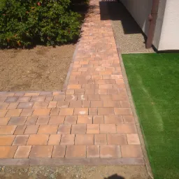 Ułożona ścieżka z ceglanej kostki brukowej prowadząca wzdłuż budynku z brązową rynną, obok sztucznej trawy i krzewu z czerwonymi owocami, cień osoby w rogu.