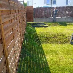Nowy drewniany płot oddzielający posesje, cień płotu na trawniku, trawnik po wertykulacji, podlewanie trawnika, schodki z kostki betonowej.