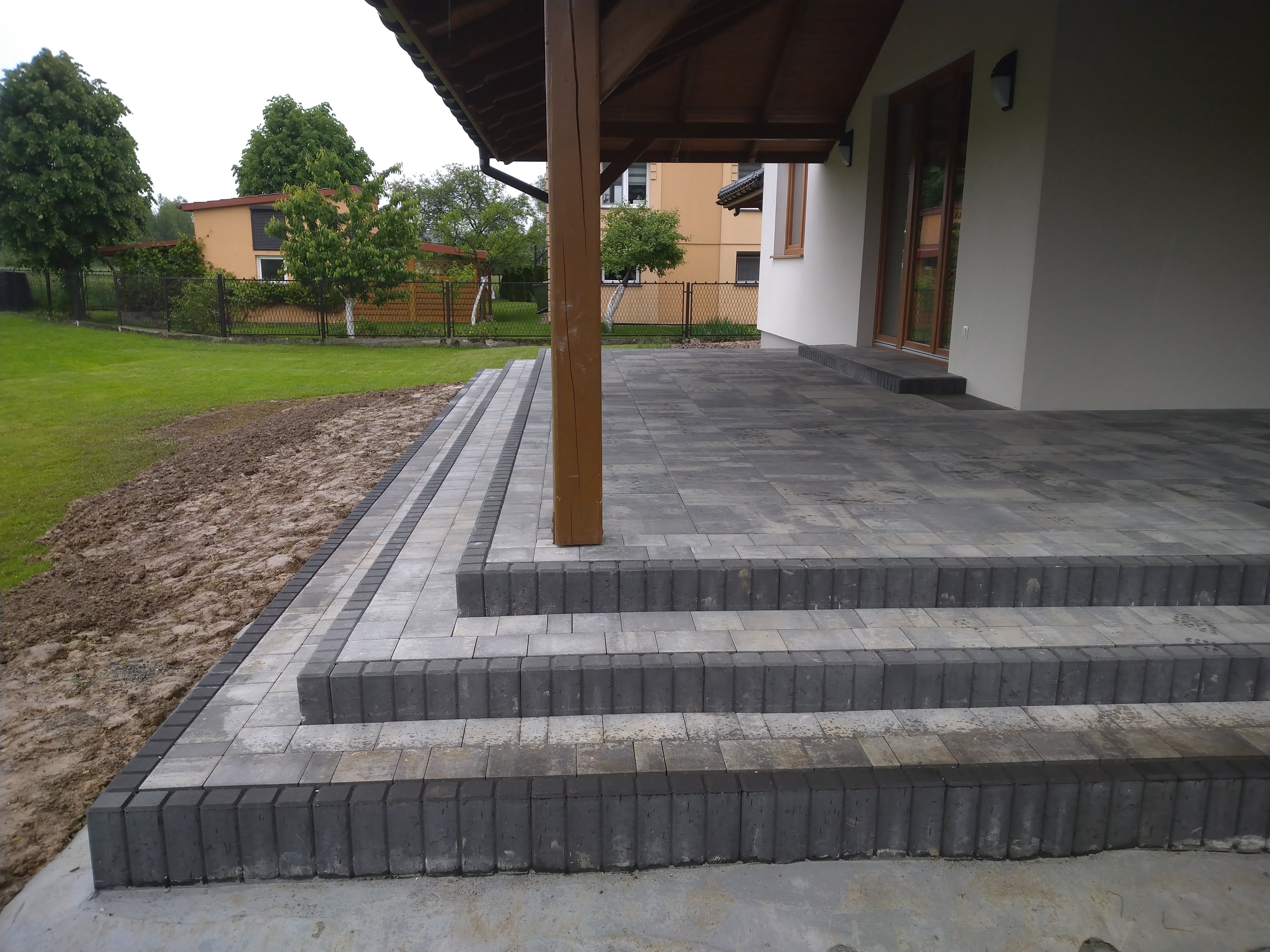 Wykończony taras z kostki brukowej w odcieniach szarości, z trzema stopniami i drewnianym słupem podtrzymującym zadaszenie, widok na trawnik i budynki w tle.