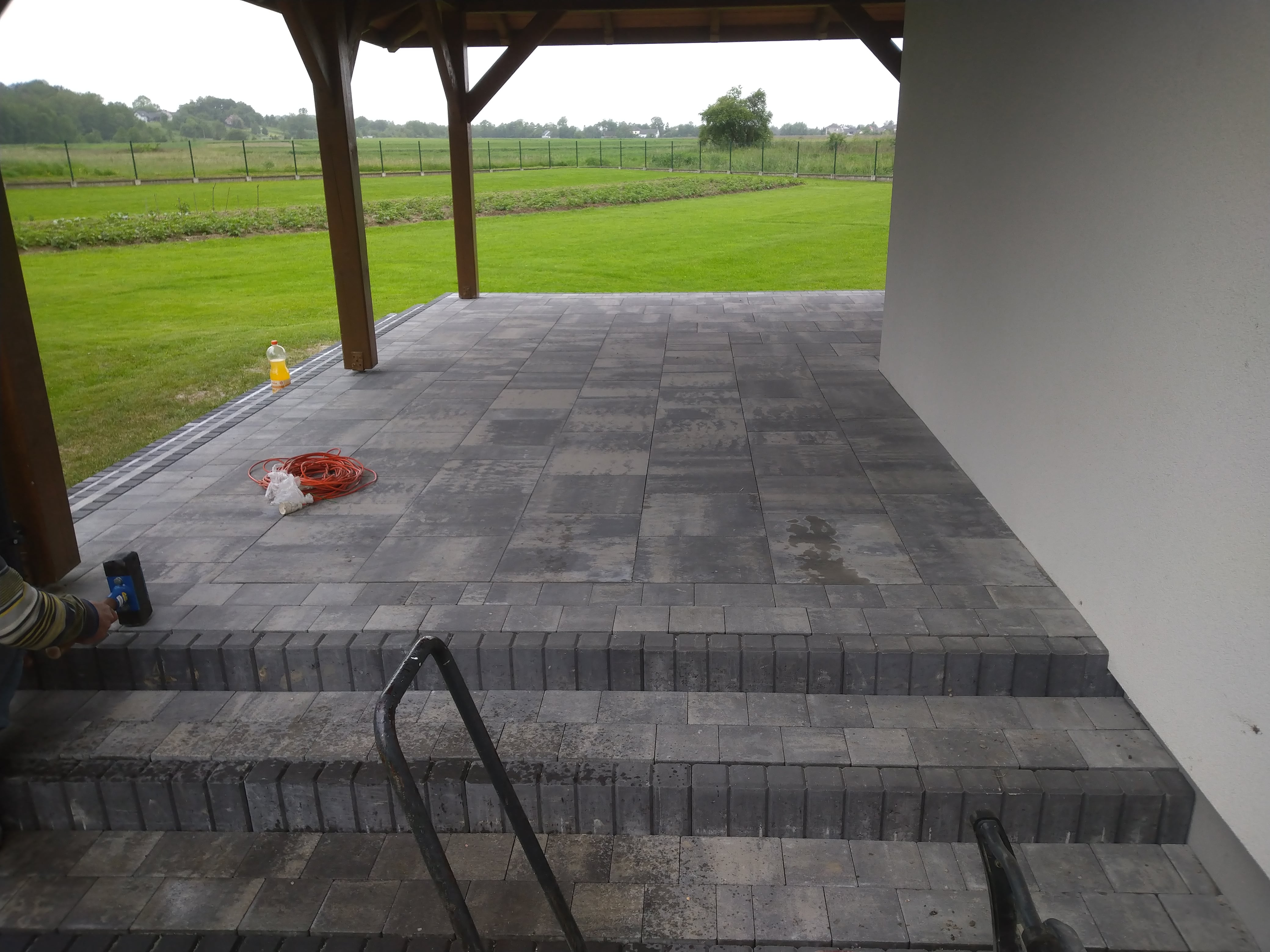 Układanie szarej kostki brukowej na tarasie z trzema schodami, widoczny pracownik z gumowym młotkiem, zwinięty pomarańczowy przedłużacz, butelka z napojem i trawnik w tle.