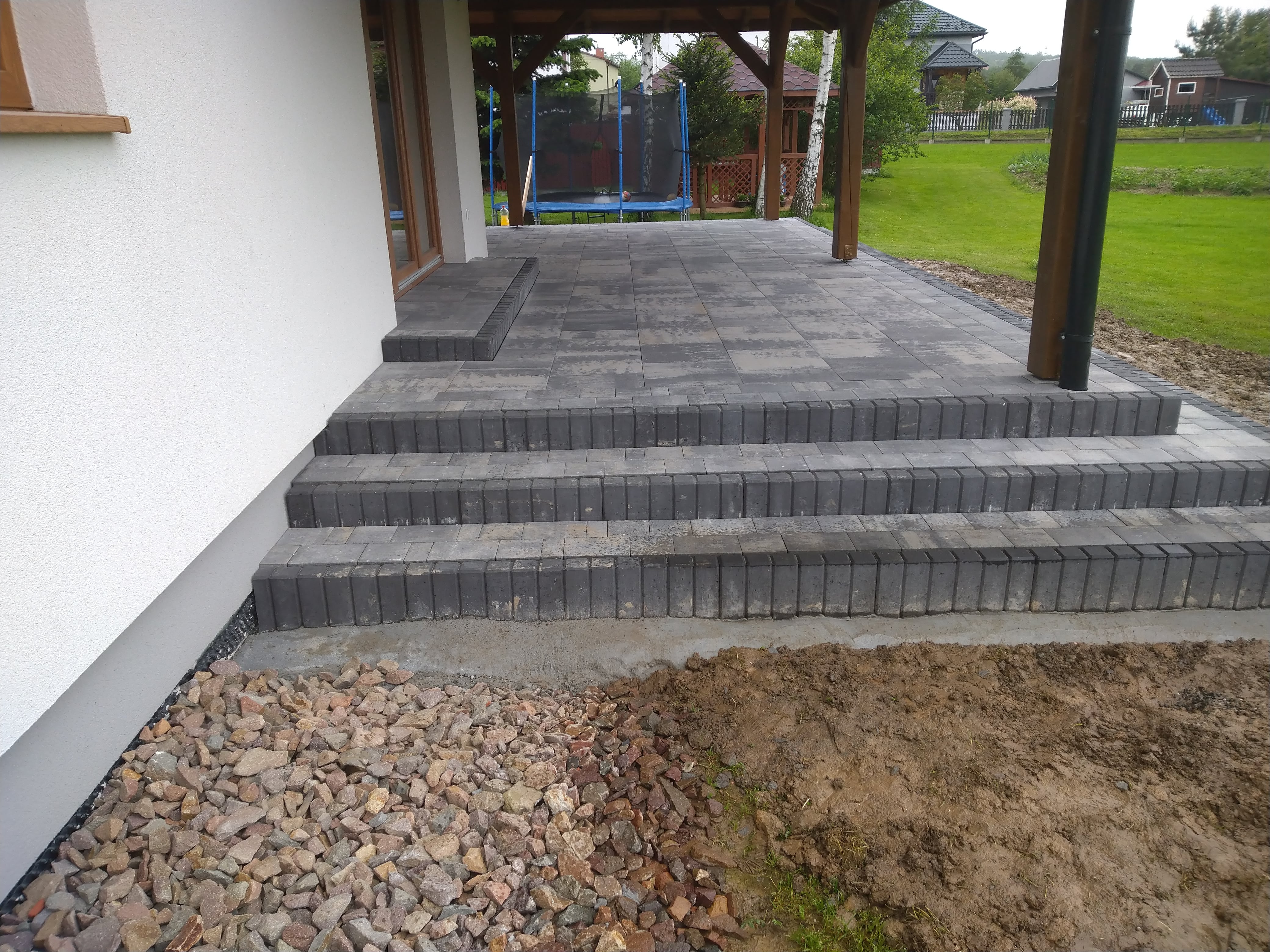 Nowo ułożona, szara kostka brukowa tworząca taras z trzema stopniami schodów przy białym domu. Widoczne kamienie ozdobne i ziemia po bokach tarasu.