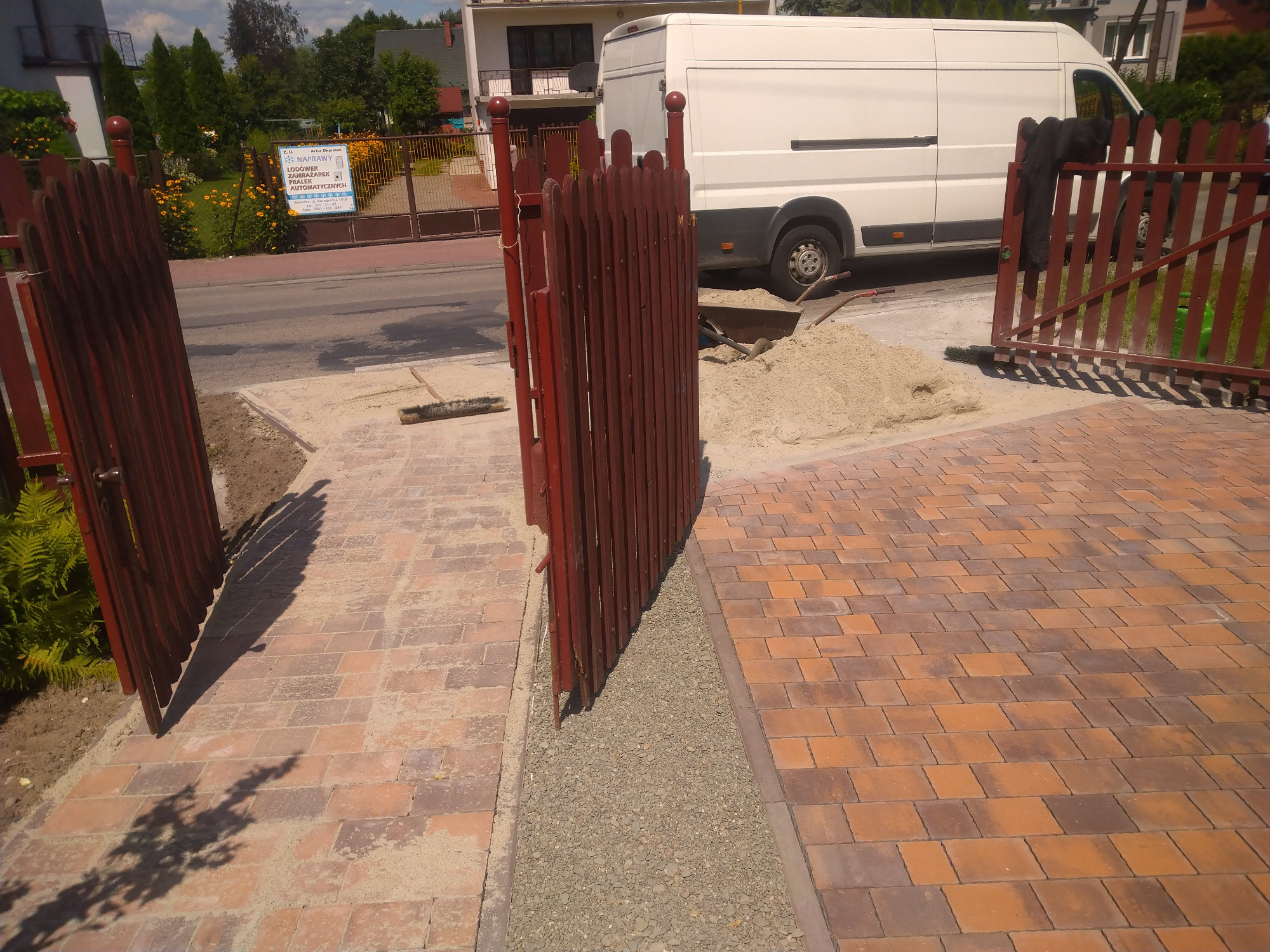 Ułożona kostka brukowa w odcieniach brązu i czerwieni, widoczna brama wejściowa z drewna oraz przygotowane podłoże z grysu, w tle biały samochód dostawczy.