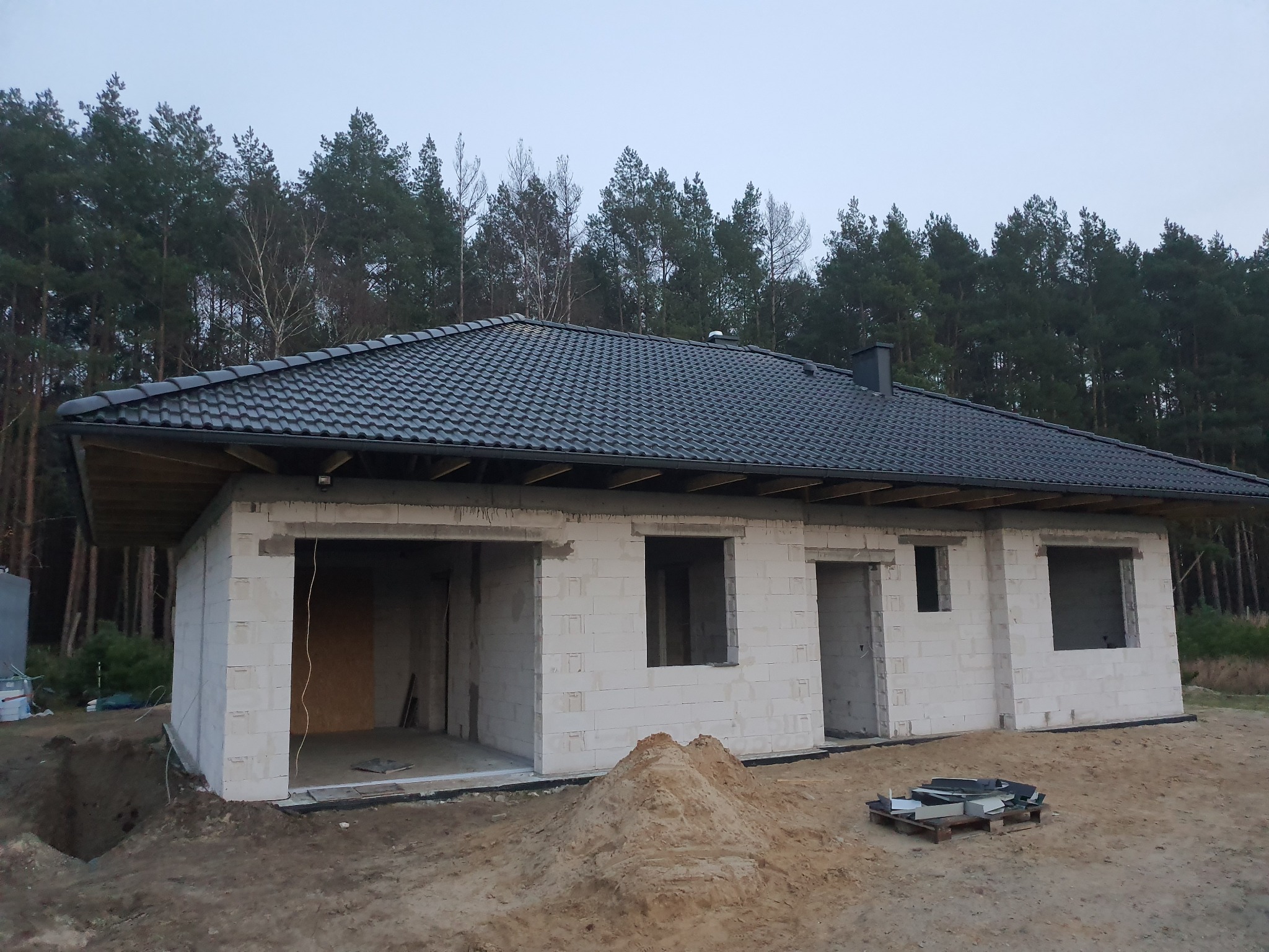 Surowy stan domu jednorodzinnego z białych bloczków, z czarnym dachem, na działce z piaskiem i lasem w tle, widoczne otwory okienne i drzwiowe, paleta z dachówkami obok.