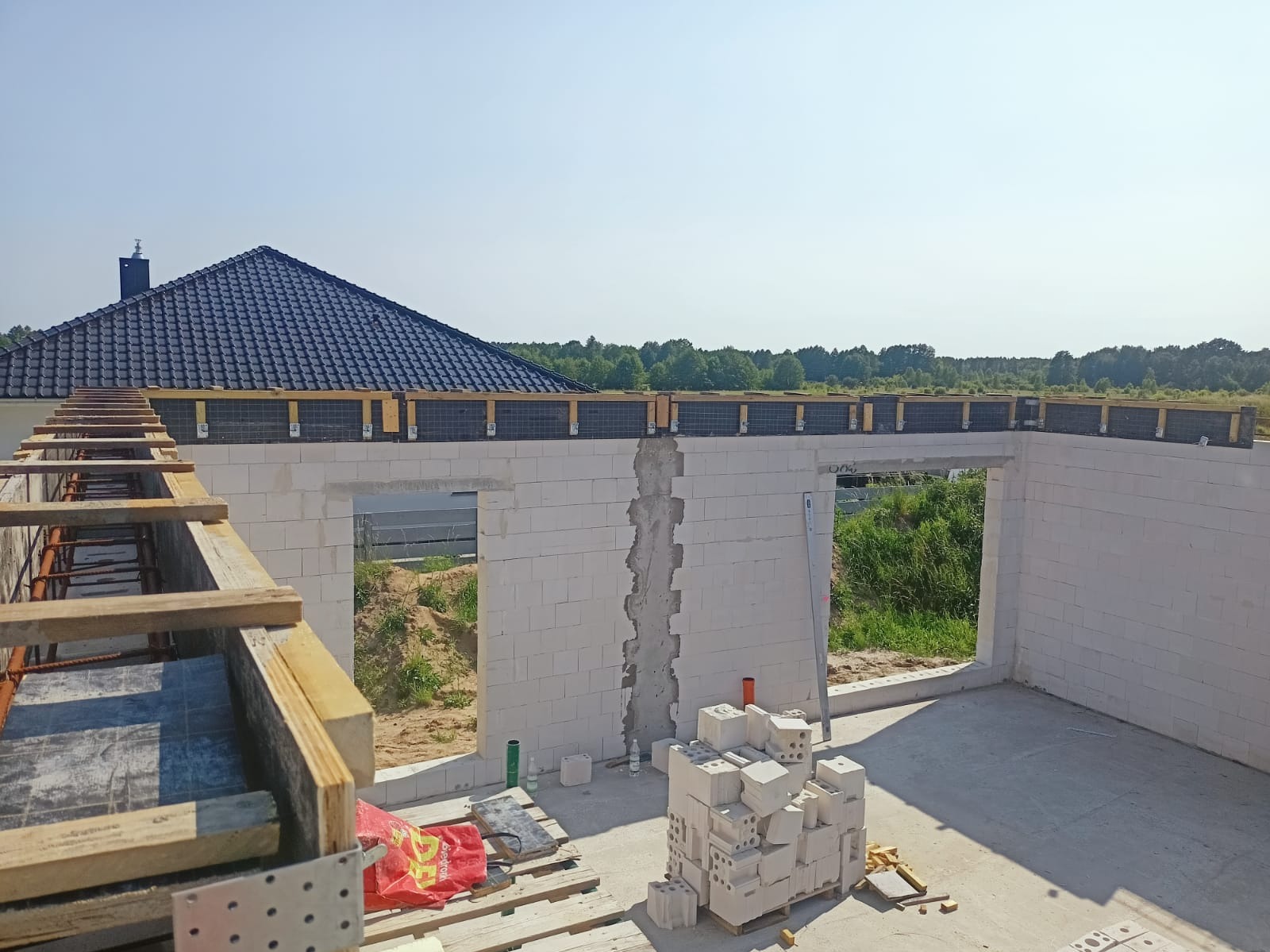 Budowa domu: ściany z białego betonu komórkowego, widoczne otwory okienne i drzwiowe, drewniane szalunki stropu. W tle dach domu i zieleń.