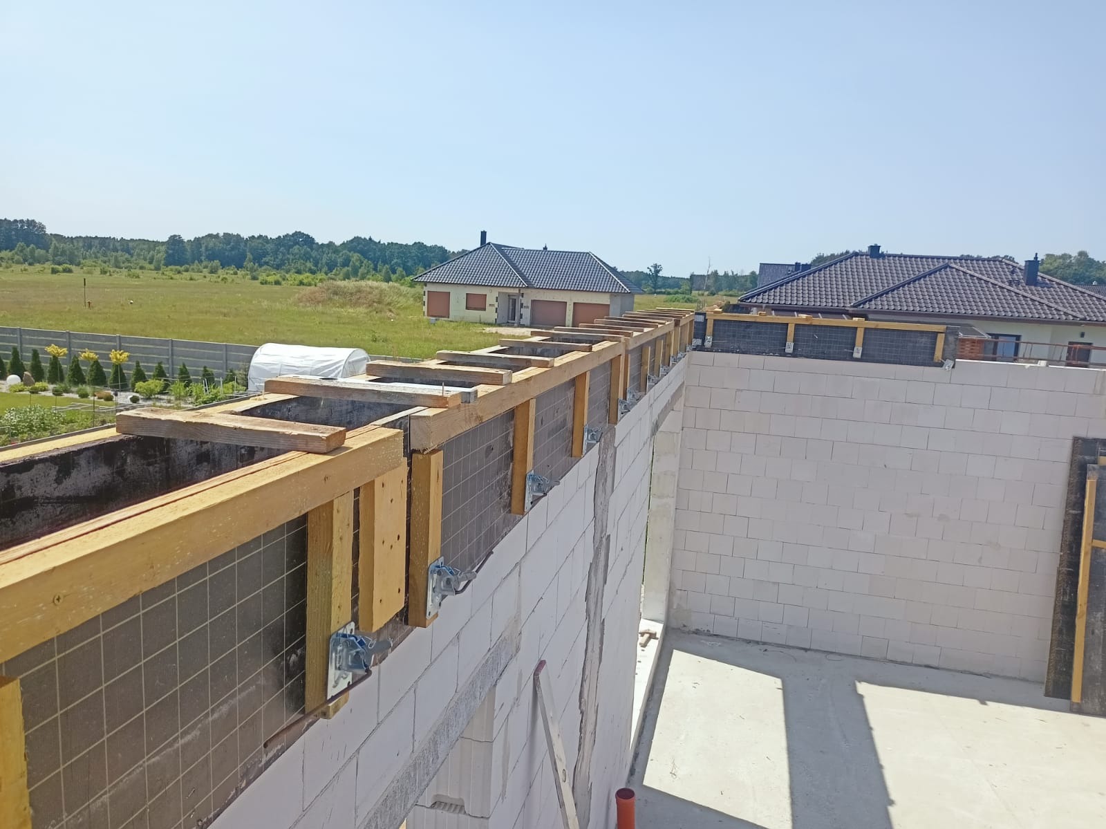 Budowa domu w toku: ściany z bloczków, drewniane szalunki, widok na okolicę. Etap murowania ścian i przygotowania do wylania stropu. Słoneczny dzień.