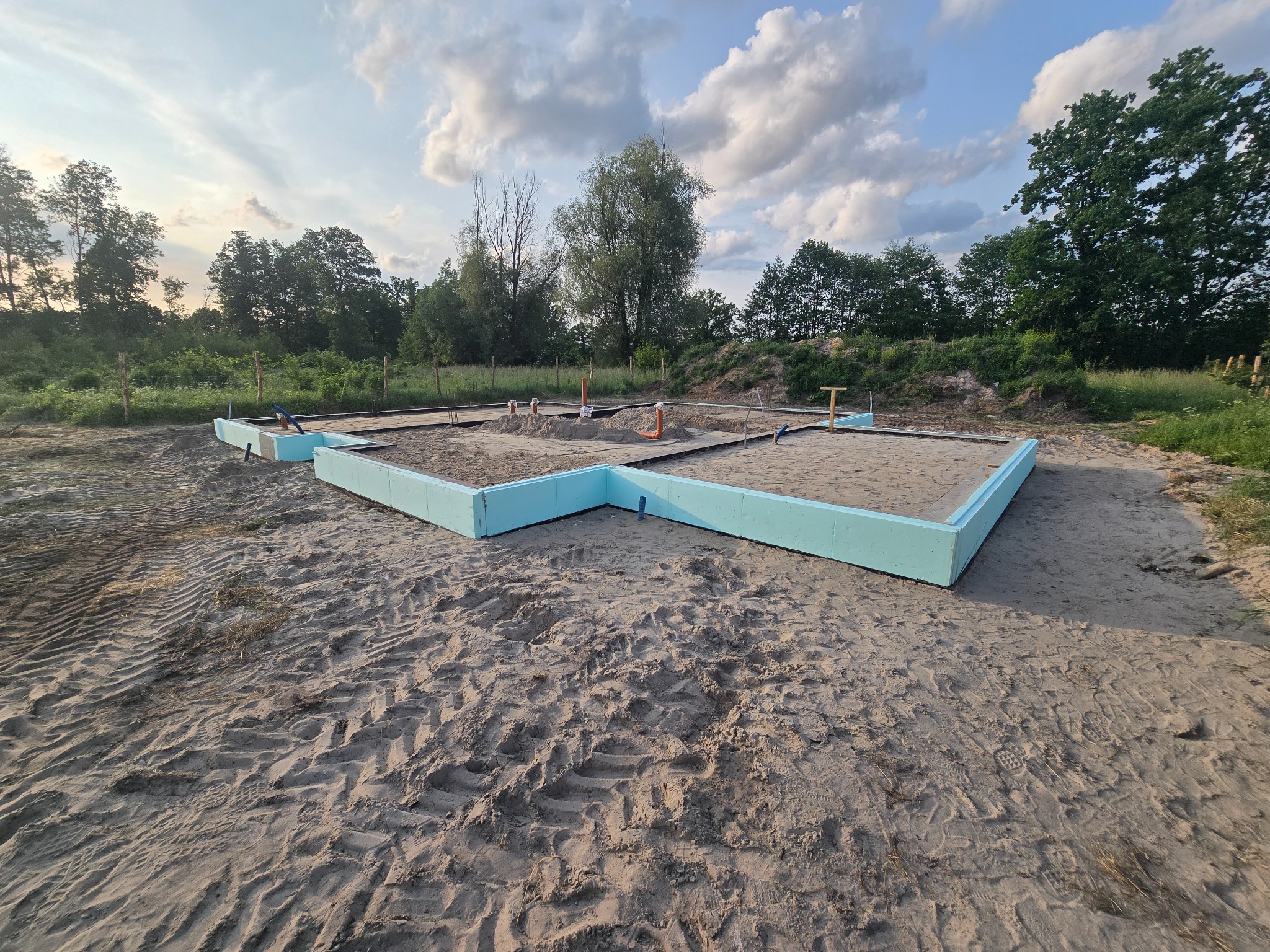 Fundamenty domu w trakcie budowy, zabezpieczone niebieskim styropianem, widoczne wystające rury instalacyjne, piaszczyste podłoże i drzewa w tle.