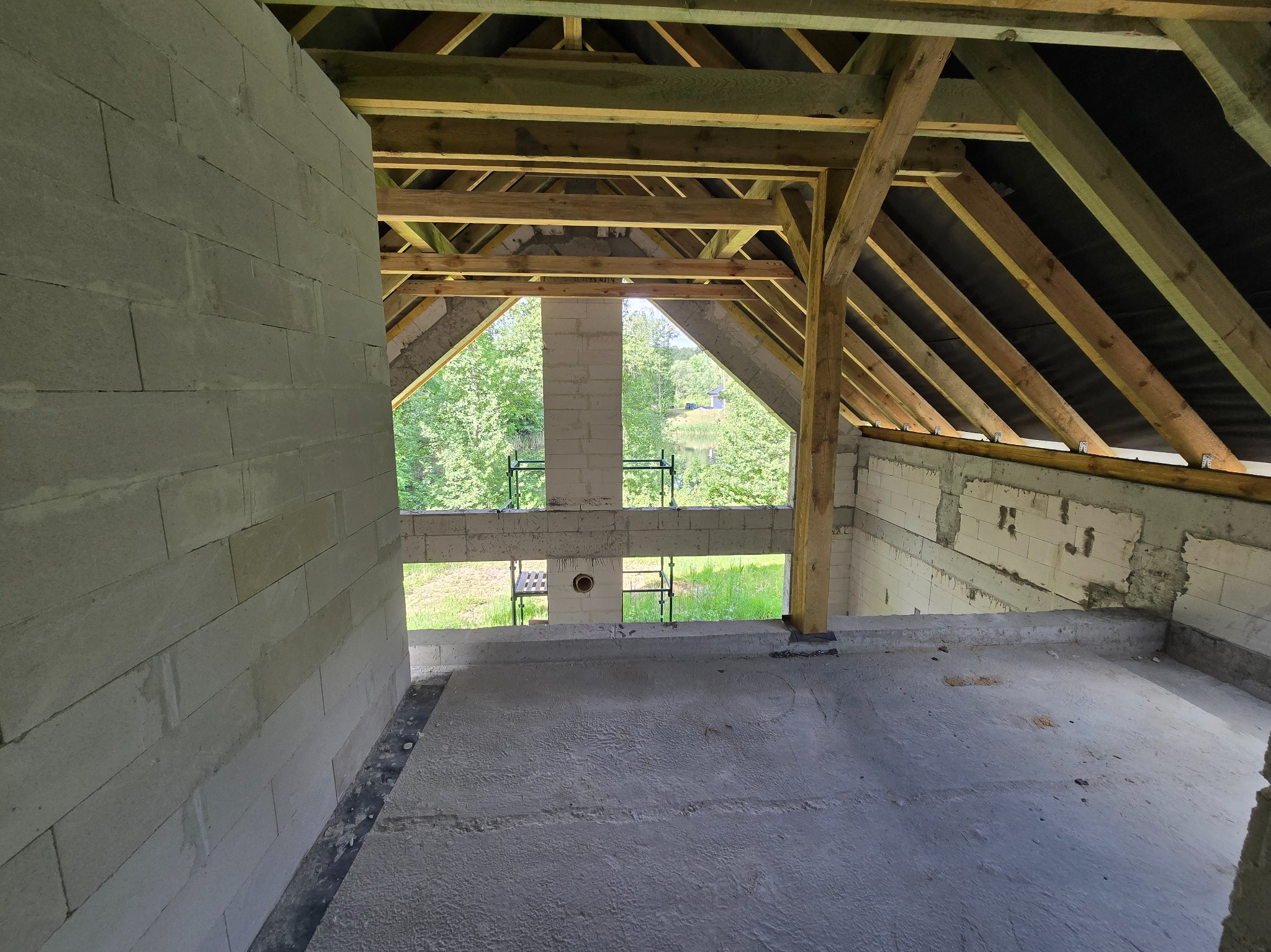 Wnętrze domu w stanie surowym otwartym: ściany z bloczków, drewniana konstrukcja dachu, widok na zieleń. Etap budowy z wylewką betonową podłogi i kominem.