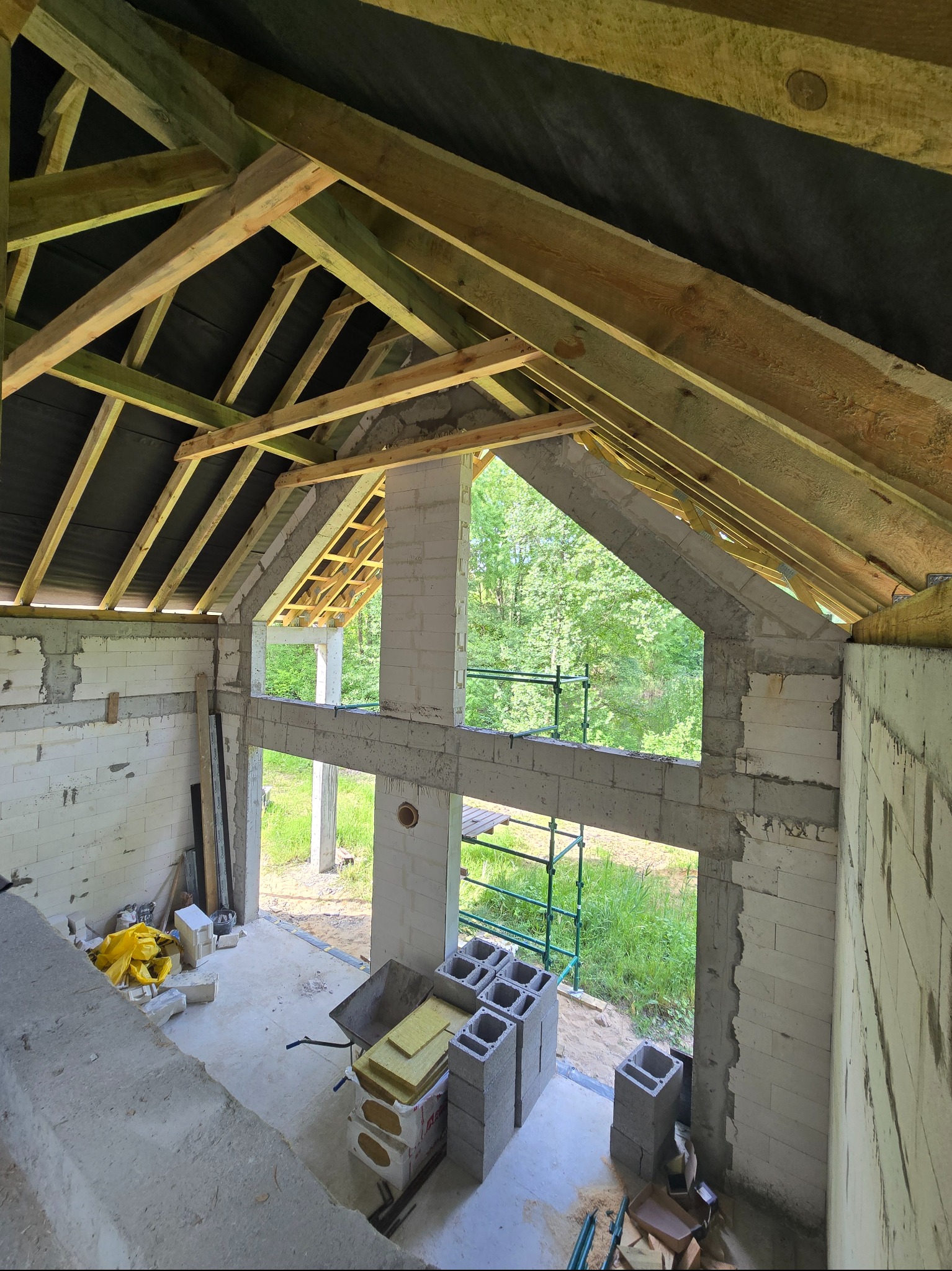 Wnętrze surowego domu w trakcie budowy: widoczne ściany z bloczków, drewniana konstrukcja dachu z membraną, okna z widokiem na zieleń, materiały budowlane i taczka.