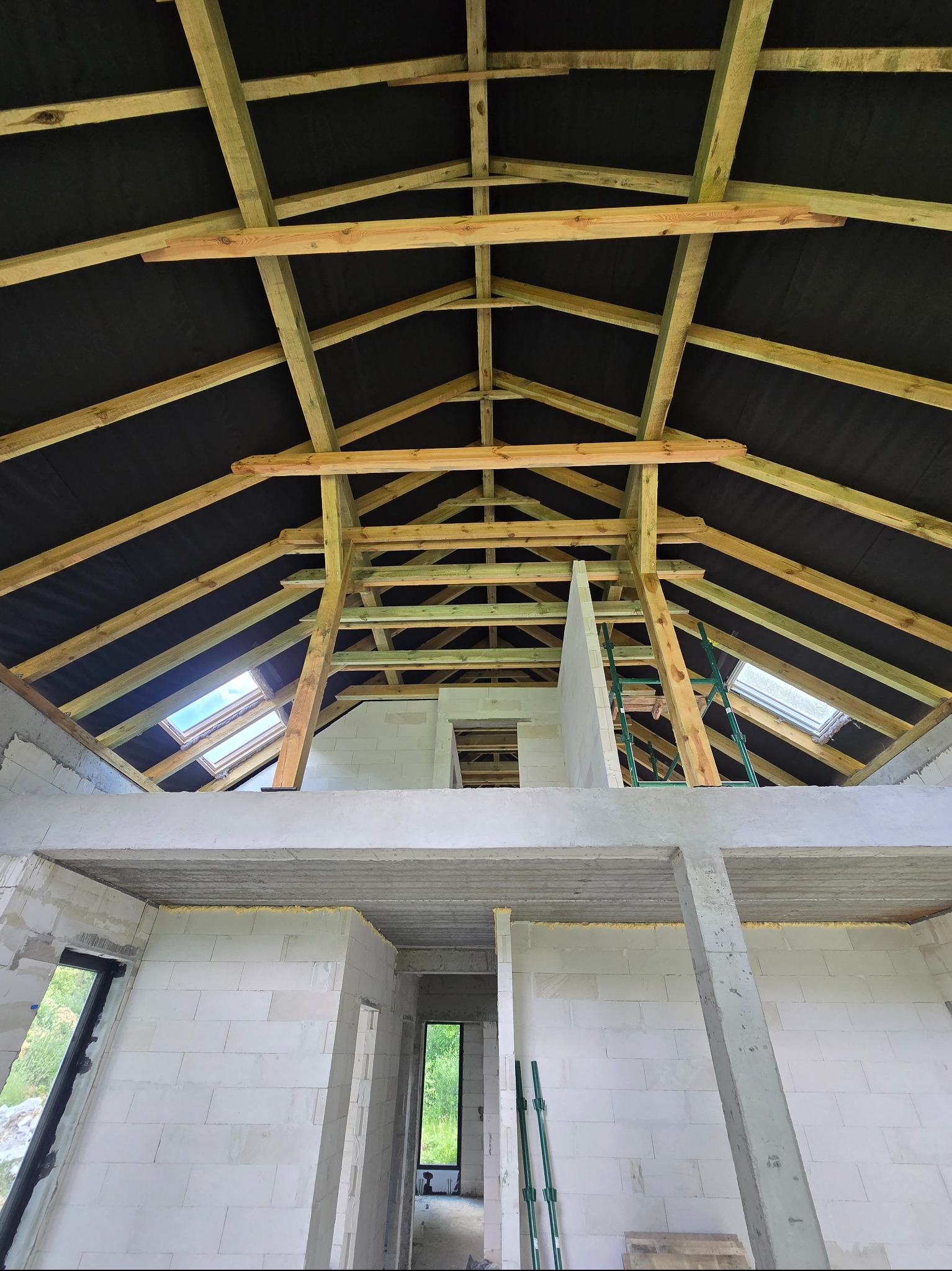 Wnętrze domu w trakcie budowy: drewniana konstrukcja dachu z czarną membraną, okna dachowe, ściany z białych bloczków, wsporniki. Widok z dołu.