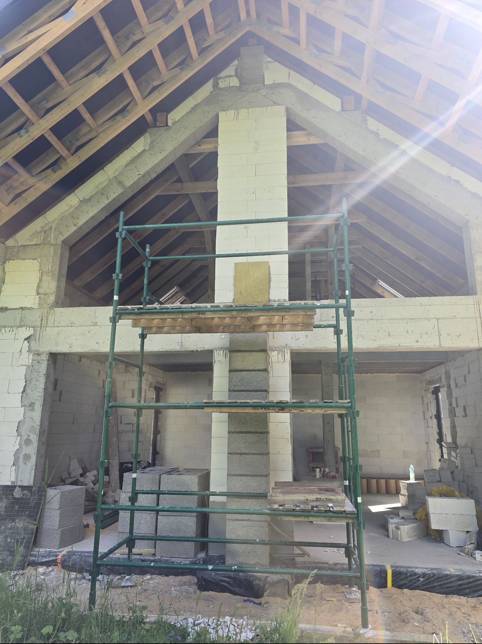 Surowy stan domu z białego pustaka, widoczny komin w trakcie budowy i drewniana konstrukcja dachu. Na pierwszym planie rusztowanie.