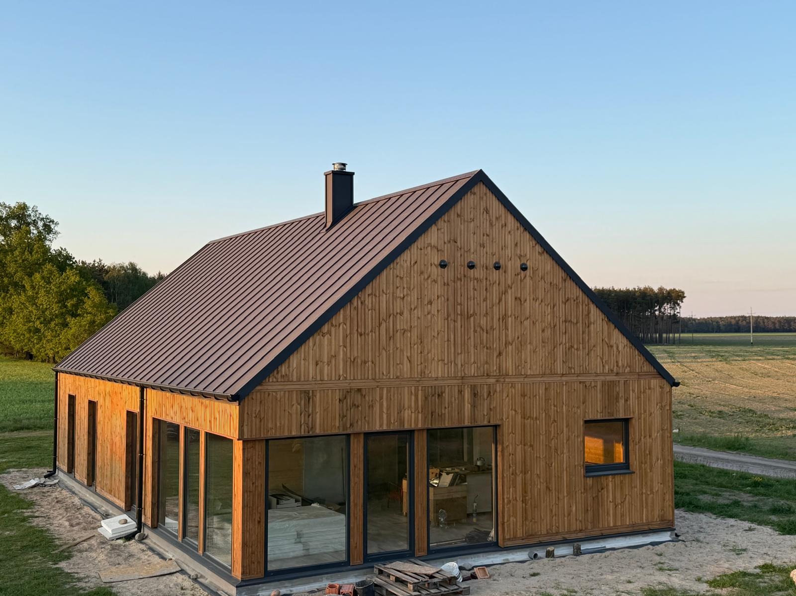 Nowoczesny dom z drewnianą elewacją i ciemnym dachem na tle zieleni i błękitnego nieba, w trakcie wykańczania. Widoczne duże okna i komin.