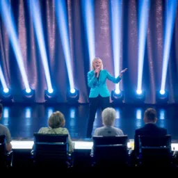 Kobieta w turkusowej marynarce śpiewa do mikrofonu na scenie oświetlonej reflektorami, przed nią siedzą osoby na krzesłach.