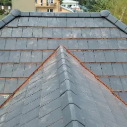 Widok z góry na szary dach z dachówki ceramicznej, z widocznymi elementami wykończeniowymi kalenicy i tłem z zielenią oraz budynkami w oddali.