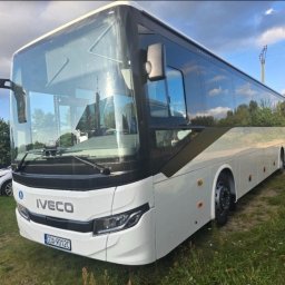 Przewozy Krajowe i Międzynarodowe KAJK TOURS K. Hingst - Biały autokar IVECO z czarnymi akcentami na trawiastym terenie, widoczny fragment zaparkowanego samochodu osobowego z przodu pojazdu.