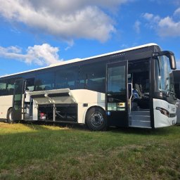 Przewozy Krajowe i Międzynarodowe KAJK TOURS K. Hingst - Nowoczesny autobus z otwartymi drzwiami i lukami bagażowymi, stojący na trawie pod błękitnym niebem. Widoczny drugi autobus w tle.