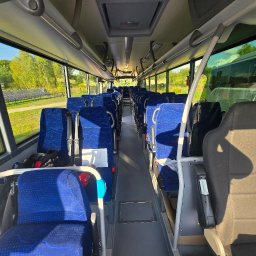 Przewozy Krajowe i Międzynarodowe KAJK TOURS K. Hingst - Wnętrze autobusu z niebieskimi fotelami, widok z przodu na puste siedzenia i okna, przez które widać zielone drzewa i panele słoneczne. Jasne oświetlenie wnętrza.