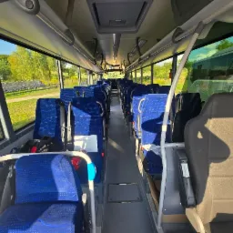 Wnętrze autobusu z niebieskimi fotelami, widok z przodu na puste siedzenia i okna, przez które widać zielone drzewa i panele słoneczne. Jasne oświetlenie wnętrza.