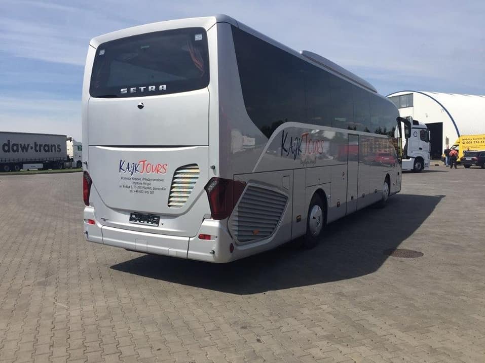 Srebrny autokar Setra z logo 'Kajk Tours' na parkingu z kostki, widok z tyłu. W tle budynek magazynowy i inne pojazdy. Słoneczny dzień.