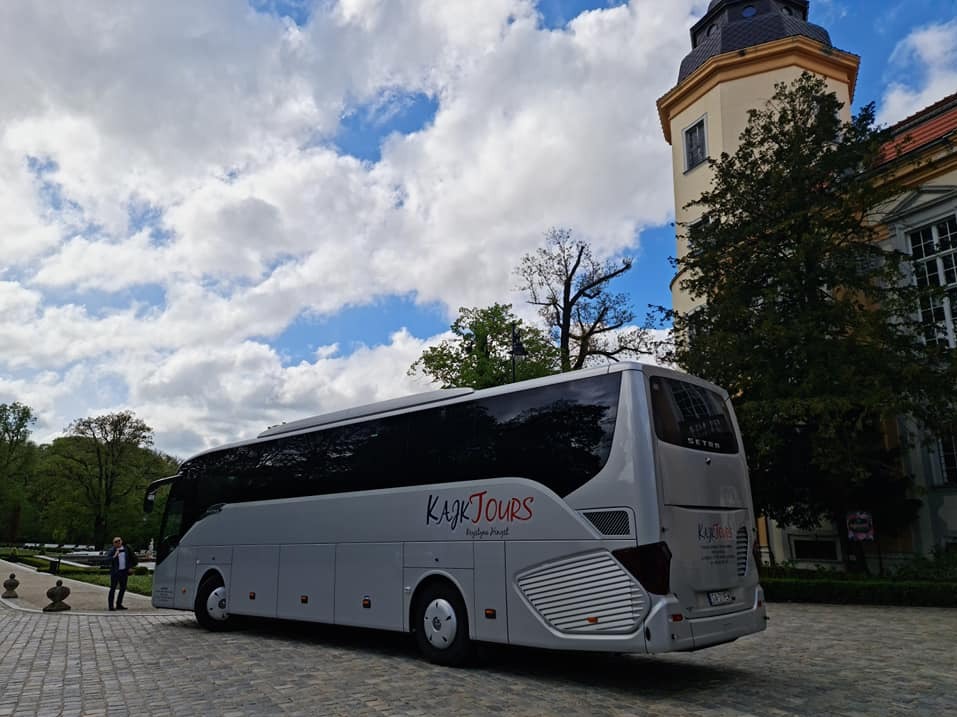 Autokar Kajk Tours z Miastka na tle pałacu i błękitnego nieba z chmurami. Widoczny mężczyzna w garniturze. Ujęcie z perspektywy chodnika.