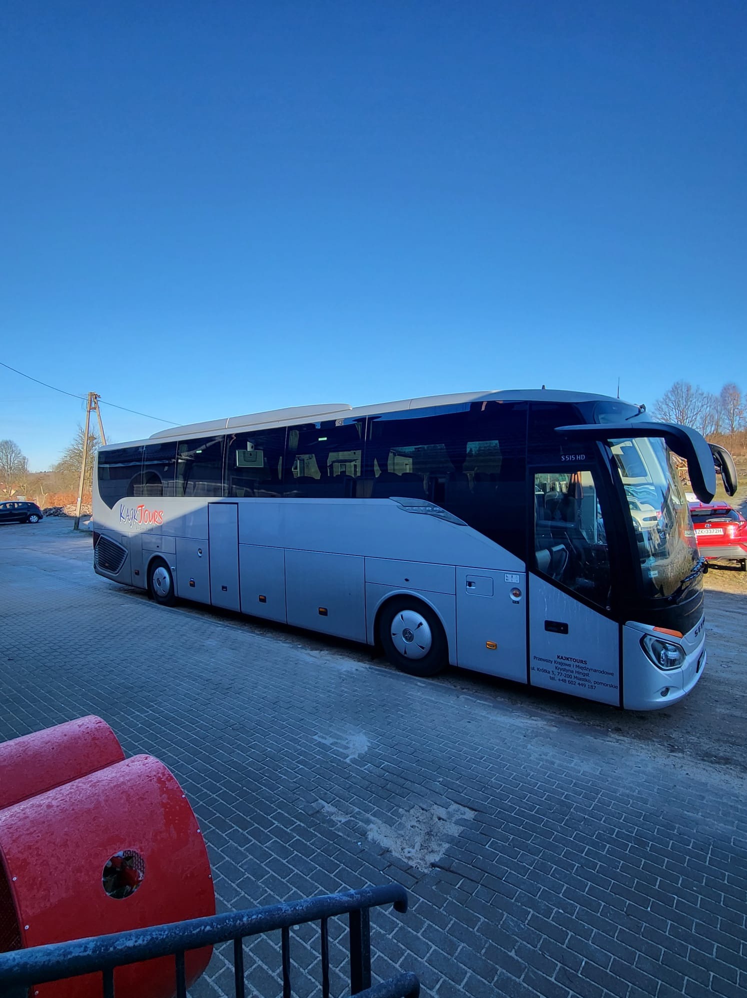 Szary autokar turystyczny z napisem 'Kajk Tours' na tle błękitnego nieba i brukowanej nawierzchni. Widoczne logo firmy i dane kontaktowe.