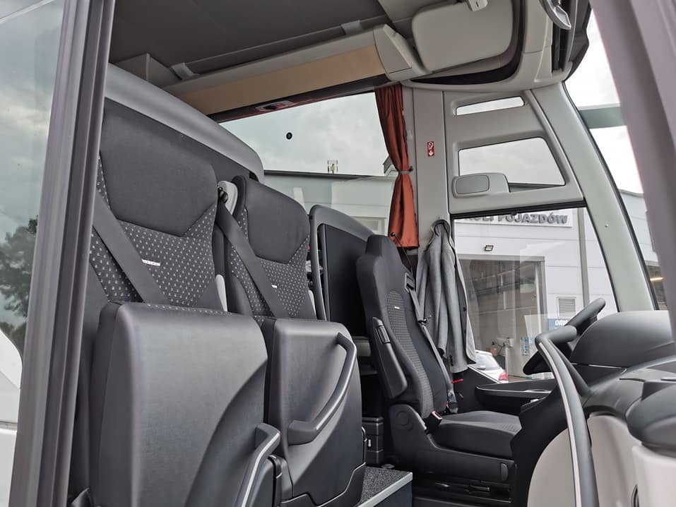 Wnętrze autobusu: czarne skórzane fotele z pasami, widok na kokpit kierowcy i budynek z napisem 'POJAZDÓW' przez okno, brązowa zasłonka przy oknie.