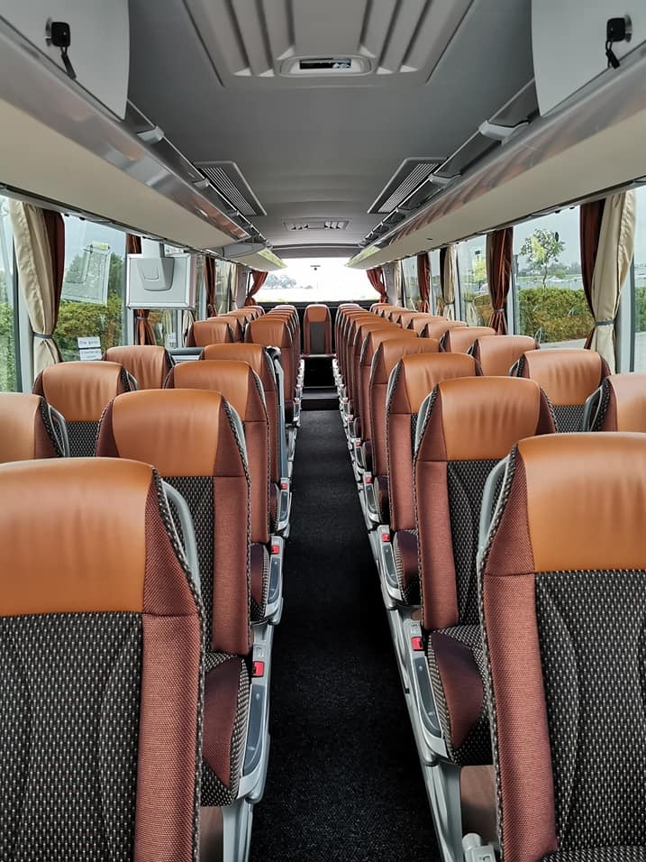 Wnętrze autobusu turystycznego: brązowe fotele z ciemnymi wstawkami, wąski korytarz, zasłony w oknach, oświetlenie sufitowe.