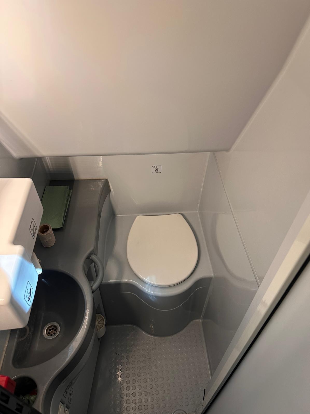 Wnętrze toalety w busie transportowym: biała muszla klozetowa, szara umywalka z dozownikiem mydła i ręcznikami, podłoga z antypoślizgową fakturą.