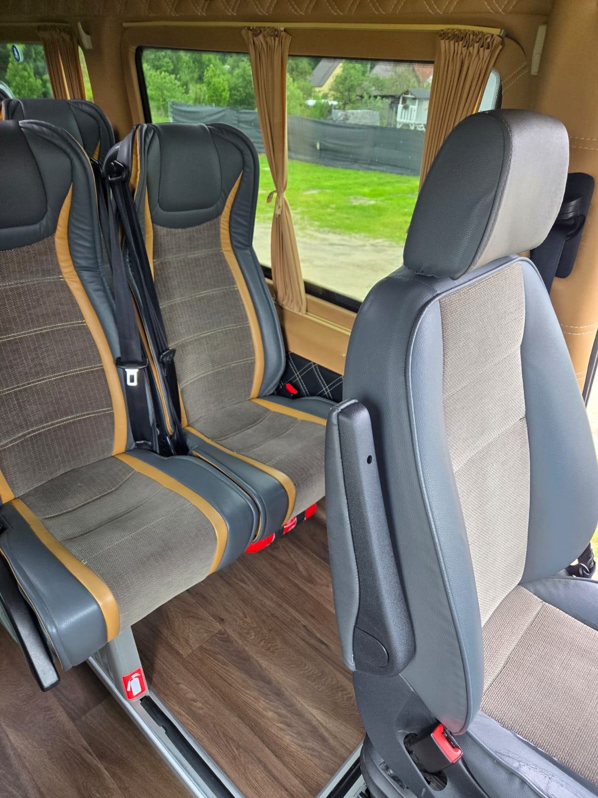 Wnętrze komfortowego busa: szare, skórzane fotele z brązowymi akcentami, pasy bezpieczeństwa, drewnopodobna podłoga, zasłony w oknach. Widok z perspektywy pasażera.