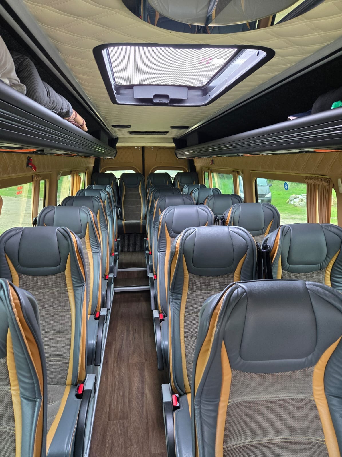 Wnętrze luksusowego busa z rzędami skórzanych foteli w odcieniach szarości i złota, drewnianą podłogą i oknami z zasłonami. Widoczny fragment luku bagażowego.