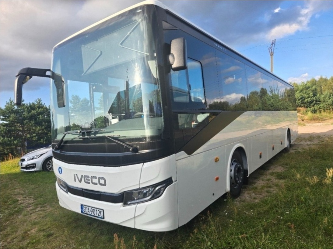 Biały autokar IVECO z czarnymi akcentami na trawiastym terenie, widoczny fragment zaparkowanego samochodu osobowego z przodu pojazdu.