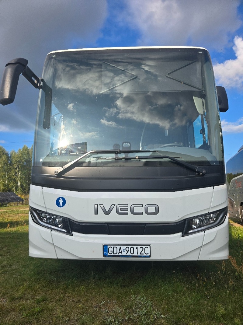Biały autobus Iveco z przodu, tablica rejestracyjna GDA 9012C, widoczne odbicie chmur w szybie, zieleń trawy i drzew w tle.
