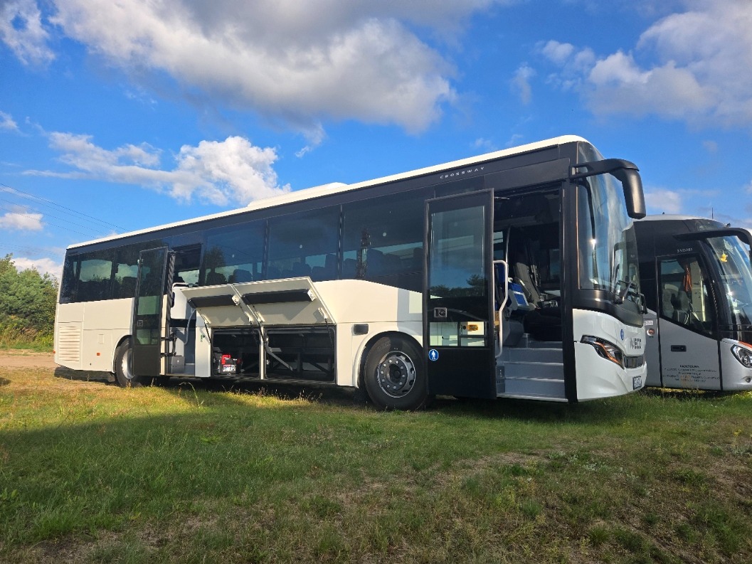 Nowoczesny autobus z otwartymi drzwiami i lukami bagażowymi, stojący na trawie pod błękitnym niebem. Widoczny drugi autobus w tle.