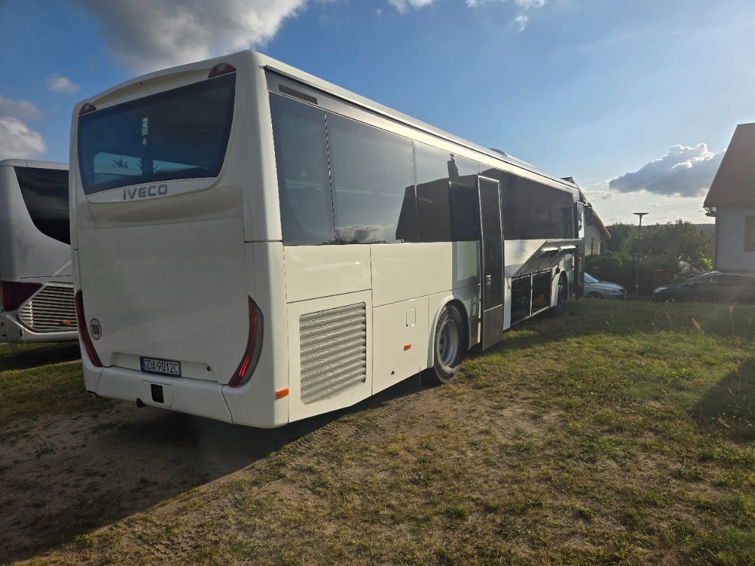 Nowoczesny, biały autokar marki IVECO z otwartymi lukami bagażowymi, zaparkowany na trawiastym terenie. Widoczny inny autokar w tle i fragment budynku. Słoneczny dzień.