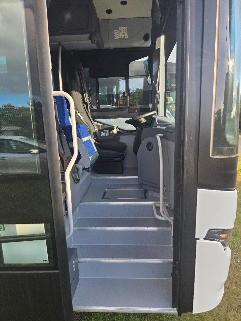 Wnętrze autobusu z widocznymi schodkami wejściowymi, siedzeniami i kokpitem kierowcy. Widok z zewnątrz przez otwarte drzwi, zieleń w tle.