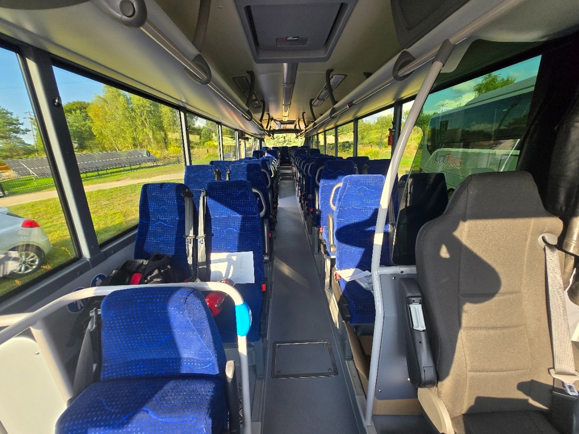 Wnętrze autobusu z niebieskimi fotelami, widok z przodu na puste siedzenia i okna, przez które widać zielone drzewa i panele słoneczne. Jasne oświetlenie wnętrza.