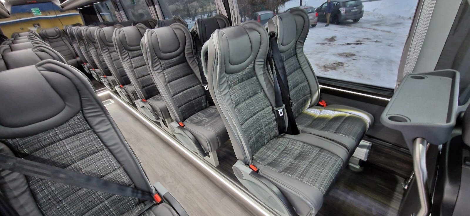 Wnętrze autobusu z rzędem szarych, tapicerowanych foteli w kratę, z pasami bezpieczeństwa i rozkładanym stolikiem. Za oknem widoczny zimowy krajobraz.