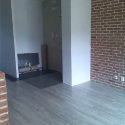 Wnętrze pokoju z panelami podłogowymi w odcieniach szarości, ceglaną ścianą i przygotowanym miejscem na kominek.