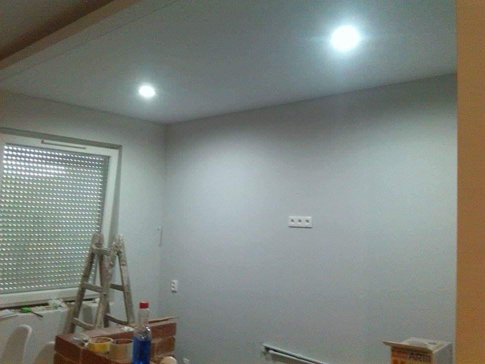 Wnętrze pokoju w trakcie malowania: drabina oparta o okno z roletami, widoczne puszki z farbą, ściany pomalowane na szaro, dwa wbudowane reflektory sufitowe.